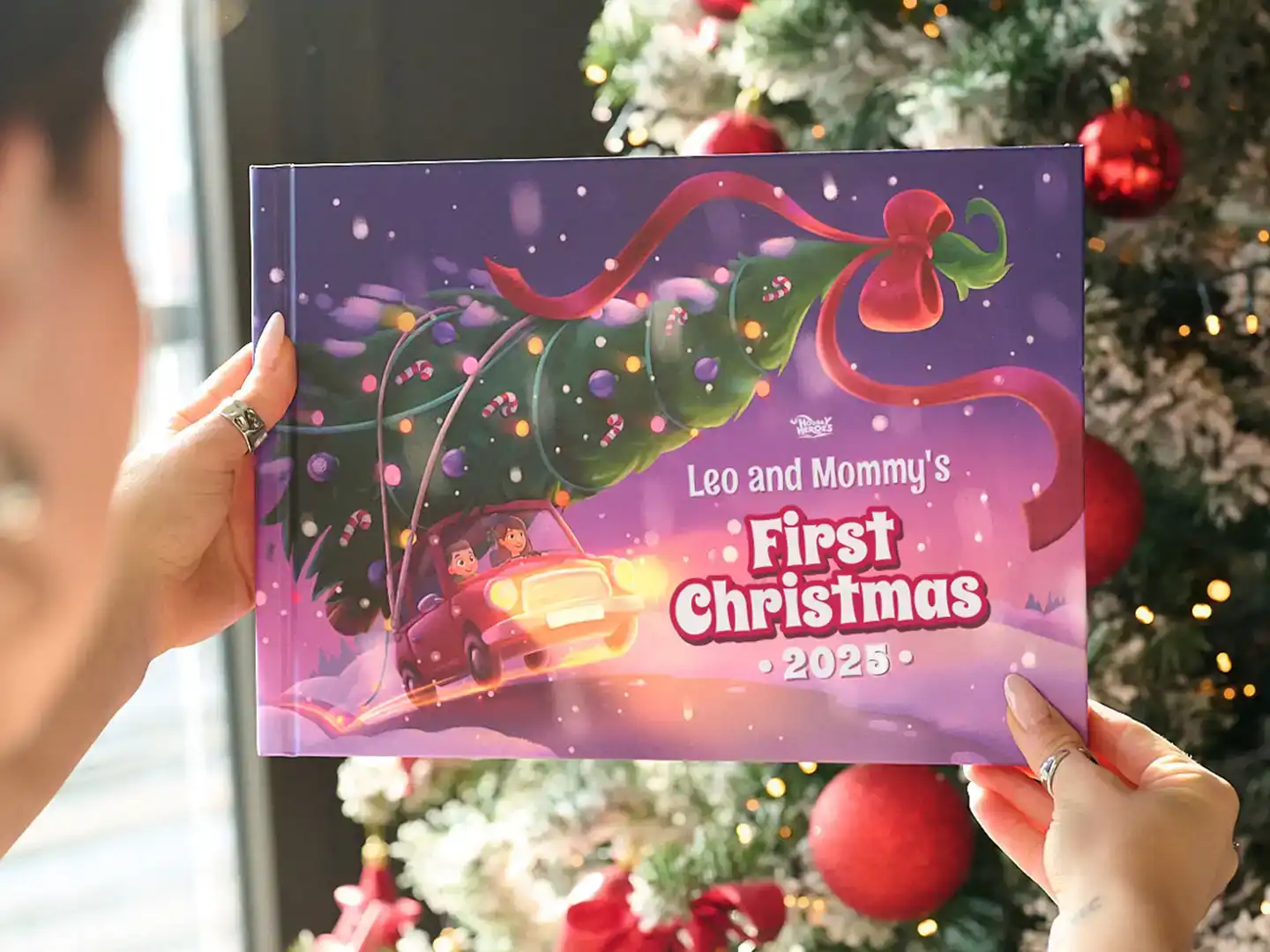 Person holding a personalized Christmas book titled “Leo and Mommy’s First Christmas 2025” in front of a decorated tree.